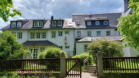 Breites Gebäude mit vielen Fenstern und Bäumen und Zaun davor